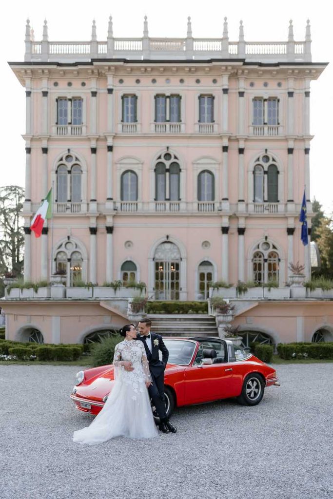 In questa foto, due sposi abbracciati davanti ad un'auto vintage di colore rosso davanti alla facciata di Villa Ponti