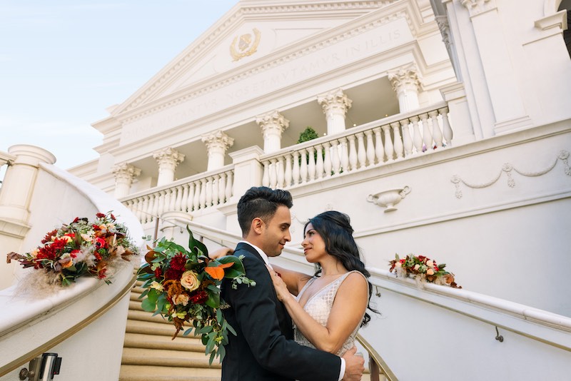 In questa foto, due sposi si abbracciano sorridenti sulla scalinata del Caesars Palace di Las Vegas. La sposa tiene un bouquet di fiori di colore rosso e arancione