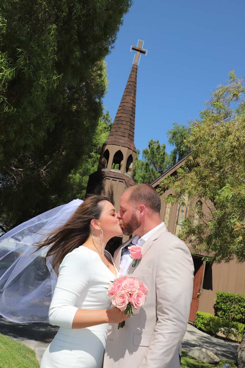 In questa foto, due sposi si baciano davanti ad una Love Chapel a Las Vegas. Lei tiene un bouquet di rose di colore rosa. Lo sposo ha una rosa nell'occhiello e indossa un abito color sabbia
