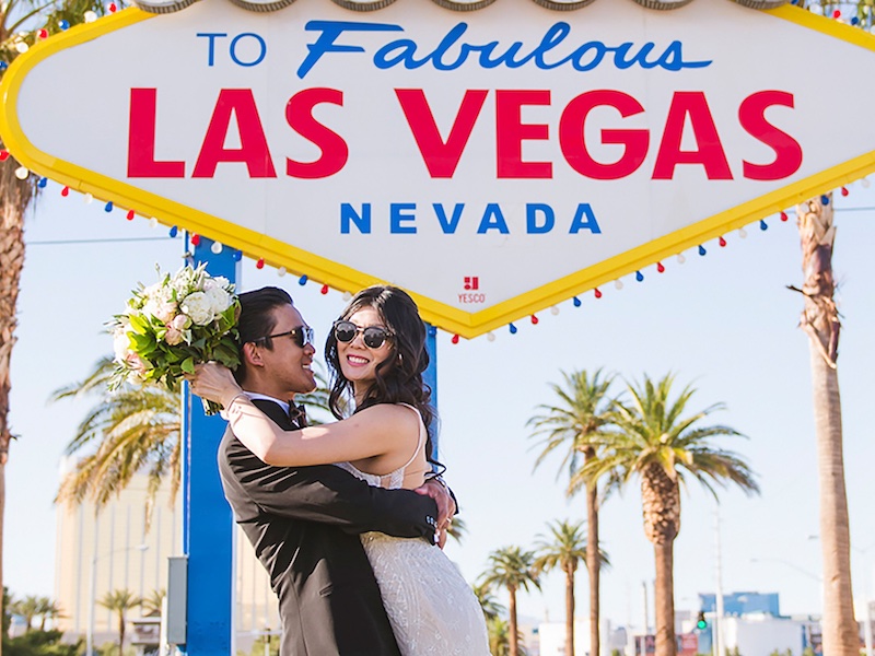 In questa foto, due sposi si abbracciano sorridenti davanti all'insegna di ingresso a Las Vegas. Entrambi indossano occhiali da sole. Le tiene un bouquet nella mano sinistra