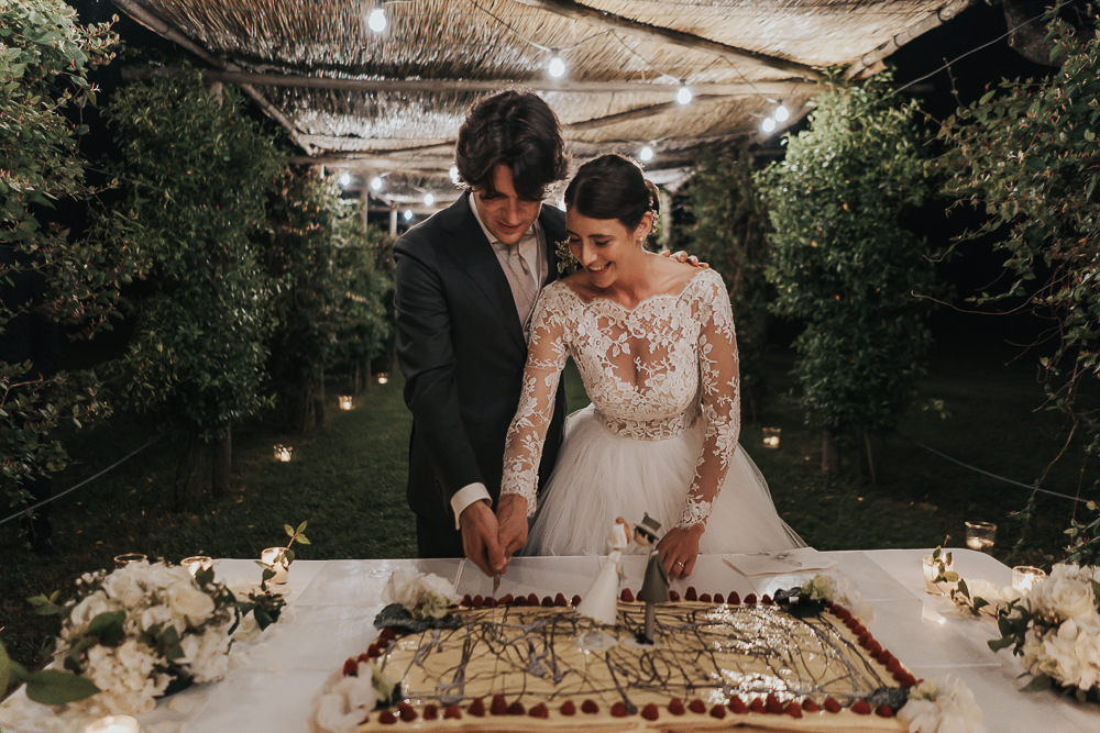 In questa foto che mostra Quale torta scegliere per il matrimonio" si vede si vede una coppia di sposi mentre taglia una torta rettangolare durante il ricevimento serale all'aperto. La sposa indossa un abito bianco con maniche lunghe in pizzo, mentre lo sposo ha un completo scuro. La torta, di colore chiaro con decorazioni scure e bordi rossi, è appoggiata su un tavolo bianco addobbato con fiori e candele, sotto un pergolato illuminato da luci soffuse.