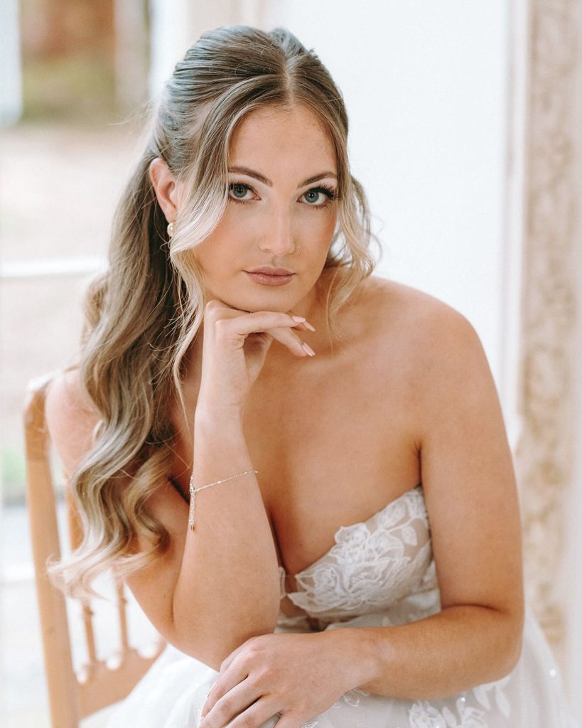 in questa foto si vede un'acconciatura sposa capelli lunghi acconciati con un mezzo raccolto con ciocche libere e morbide che incorniciano il viso. la sposa ha i capelli biondo cenere