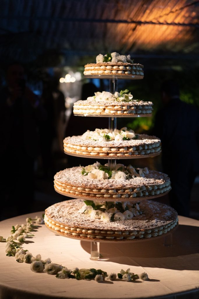 In questa foto che mostra Quale torta scegliere per il matrimonio" si vede una torta nuziale a più piani dallo stile moderno e scenografico. La torta, composta da cinque dischi sovrapposti sorretti da supporti trasparenti, è realizzata con strati di pasta sottile e crema, decorata con zucchero a velo e piccoli fiori di colore bianco. La struttura è esposta su un tavolo tondo ricoperto da una tovaglia chiara, con petali e fiori sparsi alla base. L’ambiente circostante è poco illuminato, con luci soffuse che creano un'atmosfera elegante e raccola