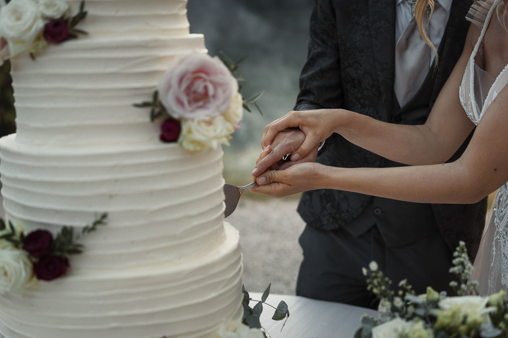 In questa foto che mostra Quale torta scegliere per il matrimonio" si vede una coppia di sposi mentre taglia insieme una torta nuziale a più piani. La torta, di colore bianco, presenta una decorazione semplice con strati orizzontali e fiori freschi applicati su vari livelli, tra cui rose di colore rosa chiaro, bordeaux e bianco. Le mani degli sposi, intrecciate, impugnano il coltello per effettuare il primo taglio. Sullo sfondo si intravedono abiti eleganti: lo sposo indossa un completo scuro, mentre la sposa ha un abito con spalline sottili e dettagli brillanti.