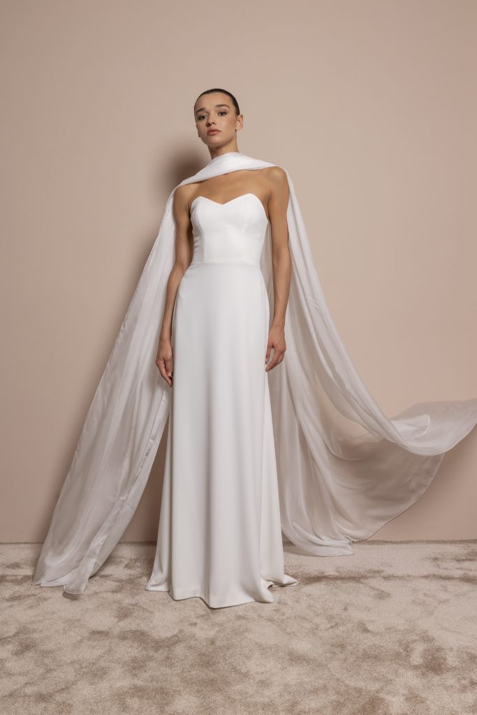 Una modella posa in piedi su un tappeto beige chiaro, davanti a una parete color cipria. Indossa un abito da sposa lungo, semplice ed elegante, di tessuto bianco liscio. Il corpetto è senza spalline, con scollo a cuore, mentre la gonna cade dritta fino ai piedi. Intorno alle spalle e al collo è drappeggiato un leggerissimo mantello in organza trasparente, che scende dietro fino al pavimento e si apre dolcemente come una coda fluttuante. La modella ha i capelli raccolti e un’espressione fiera e composta