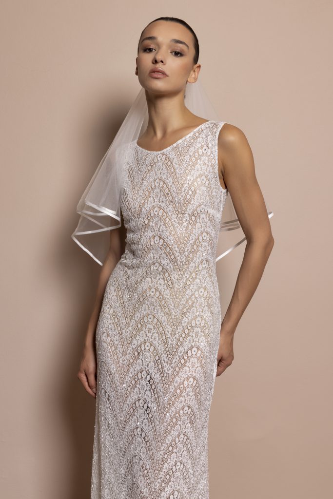 Una modella dai tratti delicati e i capelli scuri raccolti indossa un abito da sposa lungo ed elegante. Il vestito è aderente e realizzato interamente in pizzo bianco, con un raffinato motivo geometrico a ventaglio che si ripete lungo tutta la superficie. Il design senza maniche mette in risalto le spalle, mentre lo scollo è rotondo e semplice. La trasparenza del pizzo lascia intravedere un sottostrato color nude, creando un delicato contrasto. La modella indossa un velo corto in tulle con bordi rifiniti, che scende sulle spalle e dietro la schiena. Lo sfondo è uniforme color cipria, in armonia con la palette calda e raffinata dello scatto. L’espressione della modella è intensa e fiera.