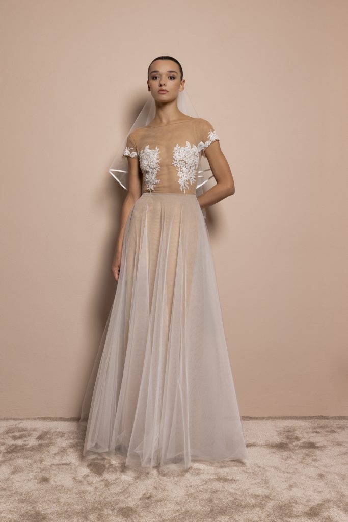 Una modella con capelli scuri raccolti indossa un abito da sposa raffinato, caratterizzato da linee pulite e dettagli delicati. Il vestito presenta un corpetto nude in tulle trasparente, con eleganti ricami floreali bianchi posizionati sul petto e lungo i fianchi. Le maniche sono corte e impreziosite anch’esse da ricami, che sembrano fiorire sulla pelle. La gonna, lunga e fluida, è composta da strati leggeri di tulle bianco che creano un effetto etereo e vaporoso. La modella indossa un velo corto in tulle con bordo definito, che scende ordinatamente dietro le spalle. Lo sfondo è di un tenue color cipria, in tono con il tappeto soffice beige ai suoi piedi. L’espressione della modella è seria e composta, in armonia con l’eleganza dell’abito.