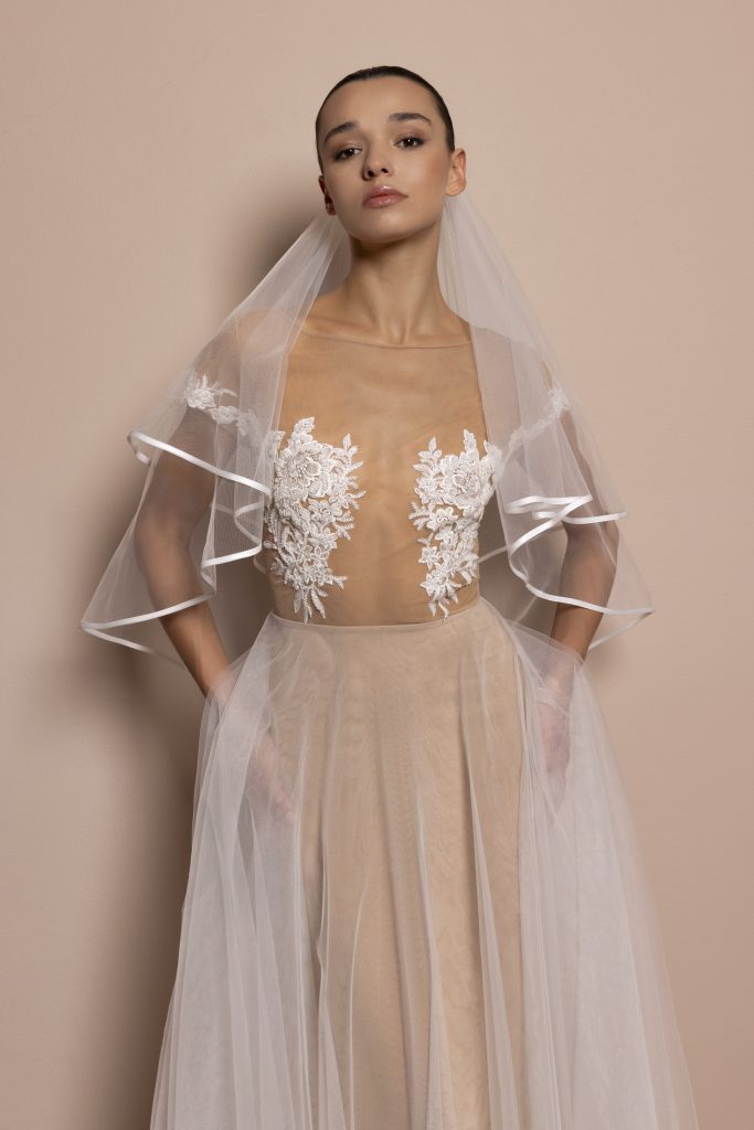 Una modella con i capelli scuri raccolti indossa un abito da sposa elegante e moderno. Il vestito è composto da un corpetto in tulle trasparente color nude, impreziosito da due ricami floreali bianchi posizionati sul petto, che donano un effetto raffinato e sensuale. La scollatura è ampia e orizzontale, con maniche in tulle leggero a volant, rifinite con un bordo bianco. La gonna è morbida, lunga e vaporosa, realizzata in strati di tulle che lasciano intravedere la base color nude sottostante. La modella indossa un velo corto in tulle bianco che cade delicatamente sulle spalle. Lo sfondo è neutro e color cipria, in armonia con i toni tenui dell’abito. L’espressione della modella è fiera e composta.