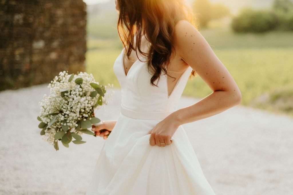 In questa foto si vede uno scatto di Ilaria&Andrea durante un momento al tramonto. La sposa è in primo piano, con un abito da sposa di colore bianco a scollo profondo e spalline larghe. Tiene con una mano un bouquet composto da fiori di colore bianco e foglie verdi, mentre con l’altra solleva leggermente il vestito. I suoi capelli castani sono sciolti e ondulati, illuminati dalla luce dorata del tramonto.