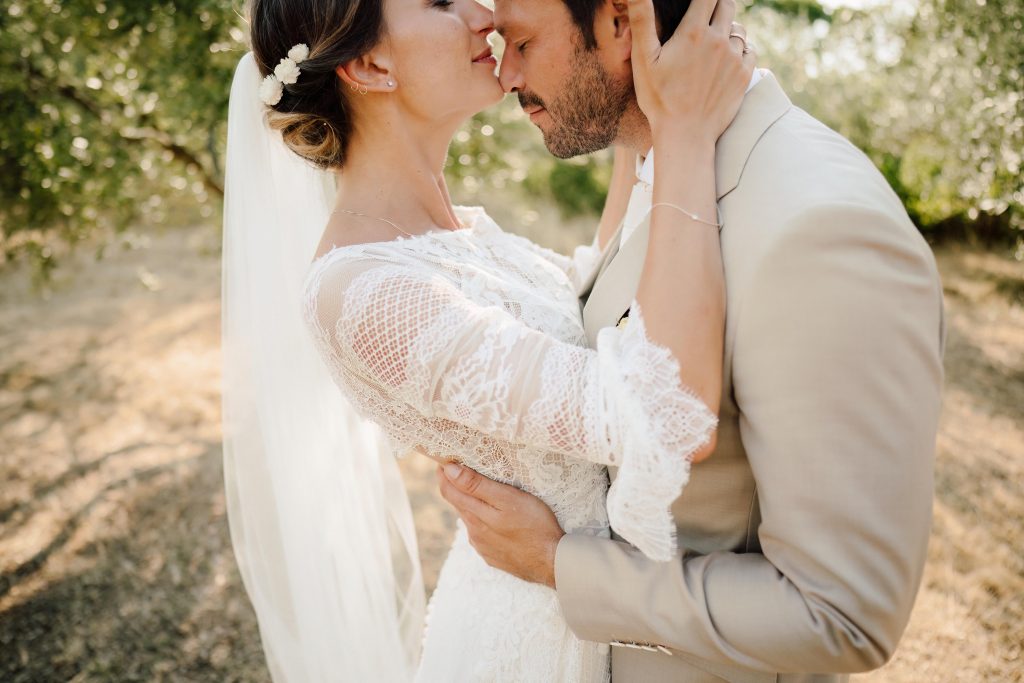 In questa foto si vede uno scatto di Ilaria&Andrea durante un momento intimo degli sposi tra gli ulivi. I due sposi si abbracciano teneramente, fronte contro fronte. Lei indossa un abito da sposa di colore bianco con maniche in pizzo e un velo lungo, mentre lui indossa un completo di colore beige chiaro. L’atmosfera è calda e romantica.