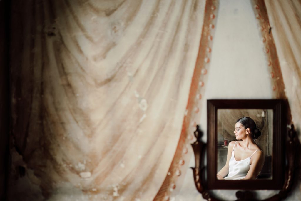 In questa foto si vede uno scatto di Ilaria&Andrea della sposa durante i preparativi del matrimonio. Riflessa in uno specchio, la sposa è seduta, con i capelli raccolti in uno chignon basso e ordinato. Indossa una canotta di colore bianco e ha un’espressione rilassata e concentrata. Sullo sfondo, un muro decorato con drappeggi pittorici in tonalità di colore beige e rosa antico.