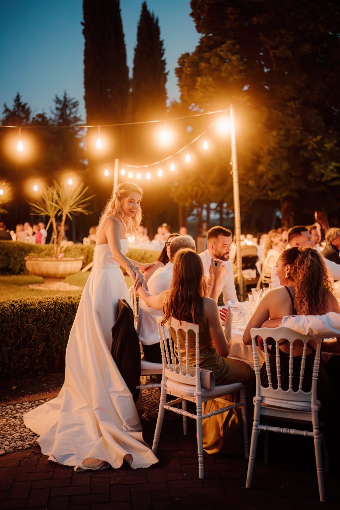 In questa foto si vede uno scatto di Ilaria&Andrea durante la cena all’aperto del loro matrimonio. La coppia è in piedi alla fine di una lunga tavolata, circondata da ospiti seduti e illuminata da una fila di lampadine sospese. Lei indossa un abito da sposa di colore bianco con lo strascico, lui è in giacca e panciotto di colore grigio scuro. L’atmosfera è calda e romantica, con luci soffuse e il cielo blu della sera.