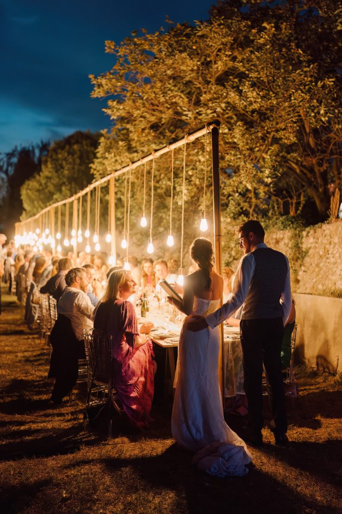 In questa foto si vede uno scatto di Ilaria&Andrea durante la cena all’aperto di un matrimonio. La scena si svolge di sera, con una lunga tavolata illuminata da tante lampadine sospese che creano un’atmosfera calda e romantica. La sposa, di spalle, indossa un abito da sposa di colore bianco e tiene in mano un libro, mentre lo sposo tiene una mano appoggiata sulla sua schiena. Gli invitati sono seduti, sorridenti, immersi nella luce soffusa.