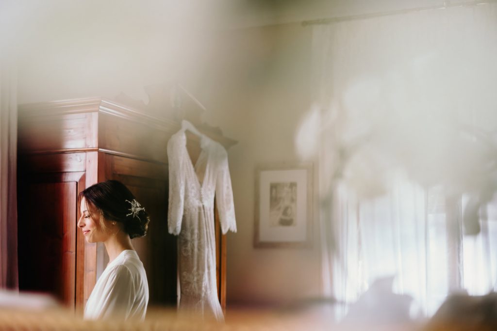 In questa foto si vede uno scatto di Ilaria&Andrea durante i preparativi del matrimonio. La sposa, di profilo, sorride e indossa una vestaglina in seta di colore bianco. I capelli sono raccolti in uno chignon basso decorato con un fermaglio floreale. Sullo sfondo, appeso a un armadio di legno scuro, si vede l’abito da sposa in pizzo, di colore bianco, illuminato da una luce soffusa.