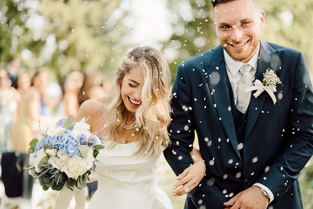 In questa foto si vede uno scatto di Ilaria&Andrea durante l’uscita dalla cerimonia. I due sorridono felici mentre vengono accolti dal lancio di petali bianchi. Lei tiene in mano un bouquet di fiori di colore bianco e azzurro, e indossa un abito da sposa con spalle scoperte. Lui indossa un abito elegante di colore blu scuro con una cravatta chiara e una boutonnière.