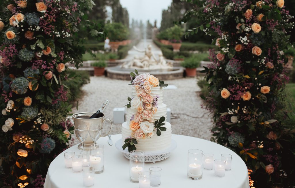 In questa foto che mostra Quale torta scegliere per il matrimonio" si vede una torta nuziale a più piani, di colore bianco, decorata con una cascata di fiori freschi nei toni del pesca, rosa cipria e lilla. La torta è appoggiata su un tavolo tondo, circondato da candele accese in bicchieri di vetro trasparente. Sullo sfondo si intravede un viale con una fontana e archi floreali ricchi di rose e ortensie