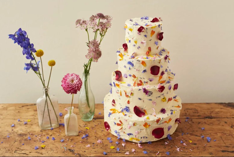 In questa foto che mostra Quale torta scegliere per il matrimonio" si vede una torta a quattro piani di colore bianco panna, decorata con petali e piccoli fiori eduli dai colori vivaci, tra cui viola, rosso, arancione, blu e giallo. La torta è poggiata su un tavolo di legno grezzo, accanto a bottigliette di vetro contenenti fiori freschi, che richiamano le tonalità della decorazione floreale della torta. L’effetto complessivo è rustico, allegro e naturale.