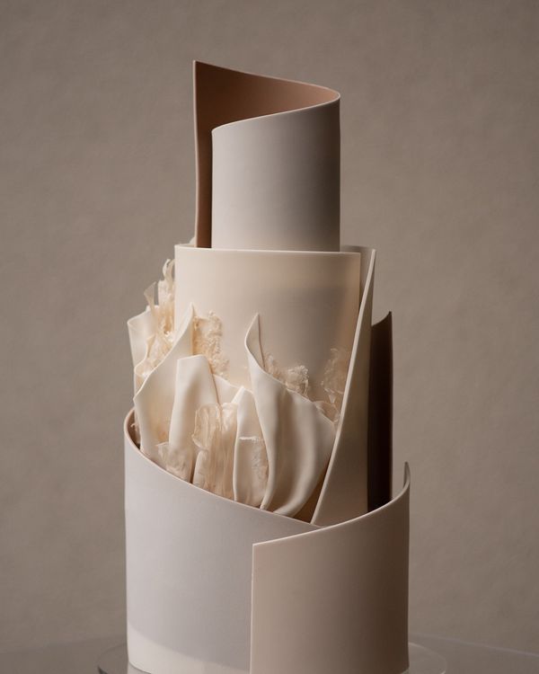 In questa foto che mostra Quale torta scegliere per il matrimonio" si vede una torta di colore avorio chiaro, moderna e scultorea, composta da più livelli rivestiti da superfici lisce e geometriche che si avvolgono a spirale. Alcuni dettagli decorativi richiamano la forma di petali arricciati, sempre nei toni del bianco e dell’avorio, dando un effetto elegante e contemporaneo.