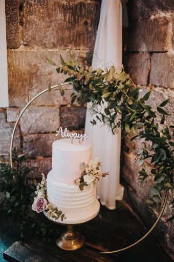 In questa foto che mostra Quale torta scegliere per il matrimonio" si vede una torta nuziale di colore bianco posizionata su un’alzata dorata. La torta è a due piani, con decorazioni floreali fresche dai toni chiari e rametti di verde. È incorniciata da un grande cerchio metallico decorato con foglie di eucalipto. Sopra la torta c’è una scritta decorativa che recita “Always”. Lo sfondo è composto da una parete in pietra e un drappo bianco.