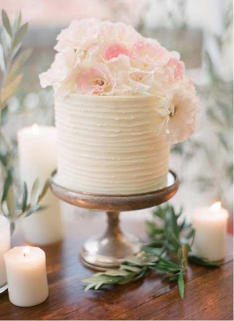 In questa foto che mostra Quale torta scegliere per il matrimonio" si vede una torta nuziale piccola e cilindrica di colore bianco, decorata con strati orizzontali di glassa. La torta è posizionata su un’alzata in metallo e adornata nella parte superiore con fiori veri di colore rosa tenue e bianco. Attorno alla base ci sono candele accese e rami d’ulivo, creando un’atmosfera romantica e naturale
