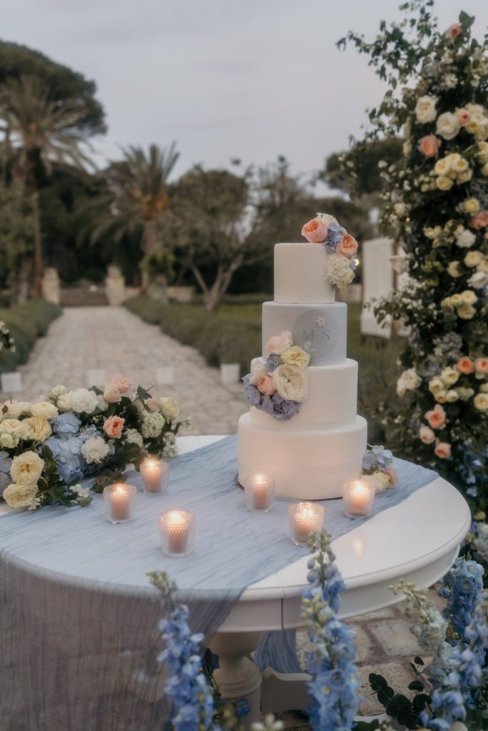 In questa foto che mostra Quale torta scegliere per il matrimonio" si vede una torta a quattro piani di colore bianco, decorata con fiori freschi nei toni del pesca, bianco e azzurro. È appoggiata su un tavolo tondo coperto da un drappo di tessuto azzurro. Intorno alla torta ci sono candele in portacandele traforati. Lo sfondo è un giardino con palme e archi floreali coordinati