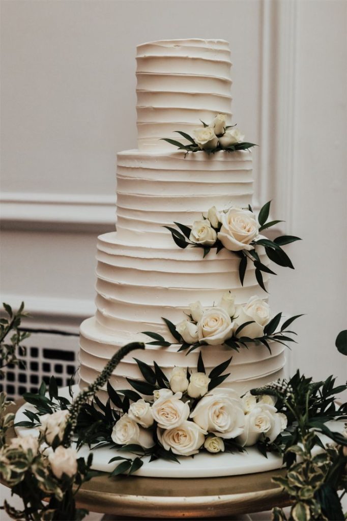 In questa foto che mostra Quale torta scegliere per il matrimonio" si vede una torta nuziale a quattro piani di colore bianco, decorata con un elegante effetto a strati orizzontali. Su ogni livello della torta sono adagiate rose fresche di colore bianco crema e foglie verdi, creando un contrasto raffinato e naturale