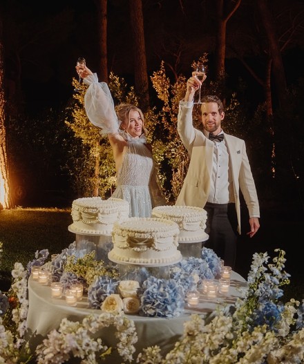 In questa foto che mostra Quale torta scegliere per il matrimonio" si vede una coppia che brinda davanti ad un tavolo allestito con decori floreali composti da rose bianche e ortensie di colore azzurro. Sul tavolo sono posizionate tre torte glassate di colore bianco