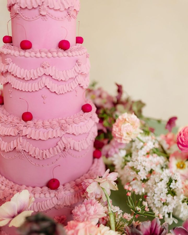 In questa foto che mostra Quale torta scegliere per il matrimonio"  si vede una torta nuziale scenografica a più piani, decorata in stile barocco con dettagli elaborati e finiture eleganti. La torta è di colore rosa intenso, con motivi a balze e ricami in pasta di zucchero. Su ogni livello sono posizionate delle ciliegie decorative di colore rosso, che creano un contrasto vivace. Alla base della torta, e in parte sullo sfondo, si vedono composizioni floreali abbondanti con fiori freschi di colore rosa, bianco e fucsia, che completano la scena con un tocco romantico