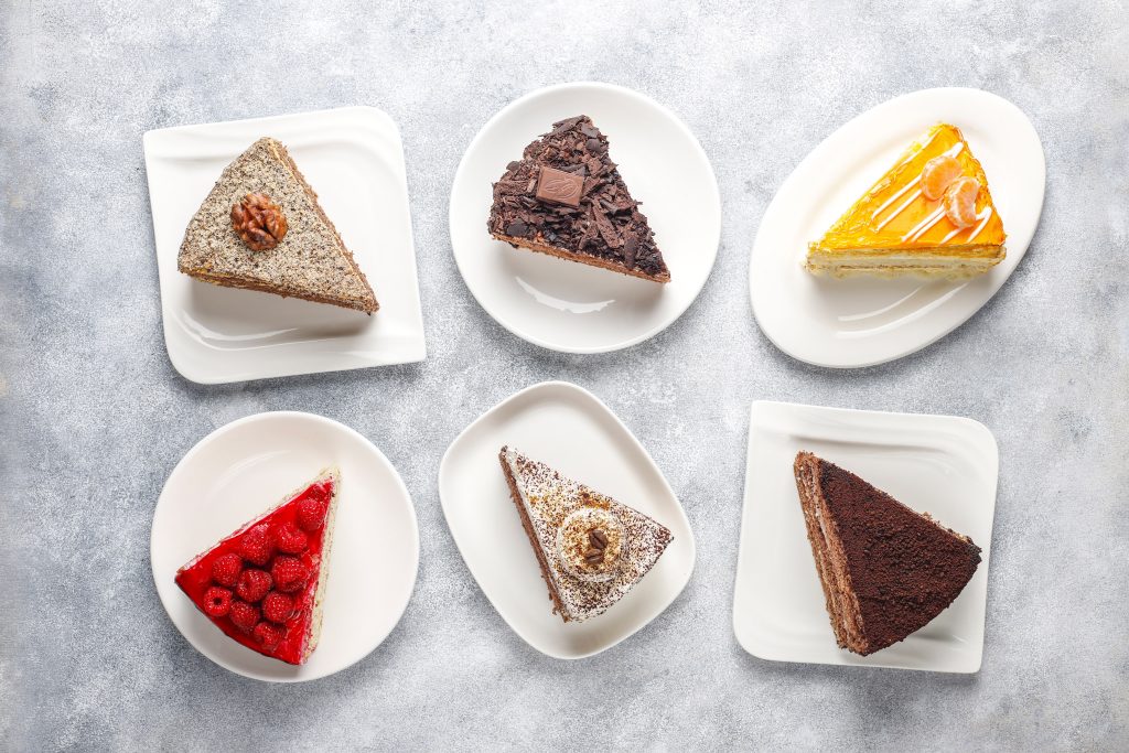 In questa foto che mostra Quale torta scegliere per il matrimonio" si vede un primo piano su sei piattini bianchi disposti su un piano chiaro, ognuno con una fetta di torta diversa. Le fette variano per colore e farcitura: una è di colore marrone scuro con crema beige, un’altra ha strati di colore rosso e bianco, un’altra ancora è bicolore con cioccolato e crema rosa, mentre le altre due mostrano farciture chiare con tocchi di colore rosso o scuro. In primo piano una fetta di torta al cioccolato di colore marrone è accompagnata da una forchetta argentata.