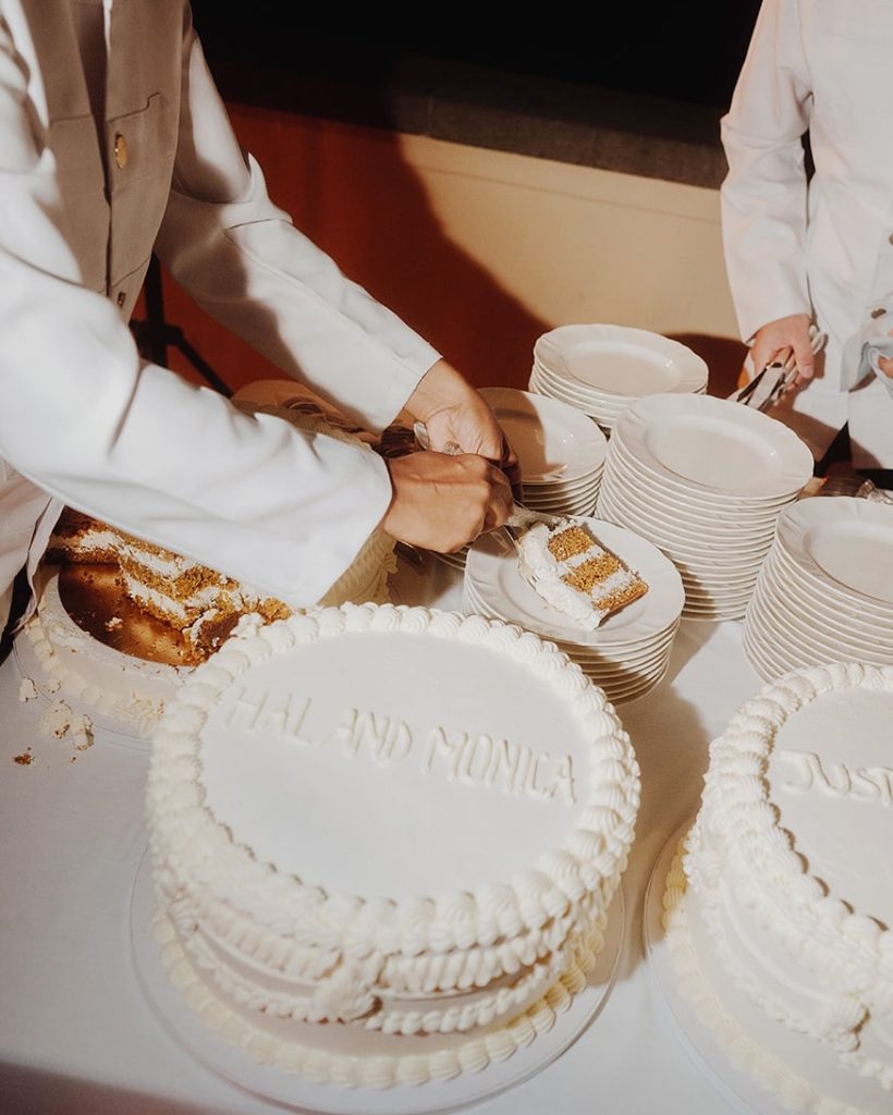 In questa foto che mostra Quale torta scegliere per il matrimonio" si vede una torta nuziale di colore bianco, decorata con semplici bordature a onde e la scritta “HAL AND MONICA” in rilievo sulla parte superiore. La torta è stata già tagliata, e una persona in divisa bianca sta servendo una fetta su un piattino, vicino a una pila di piatti pronti per gli invitati. Sul tavolo sono presenti altre torte simili, ciascuna personalizzata, in un contesto elegante e ordinato.