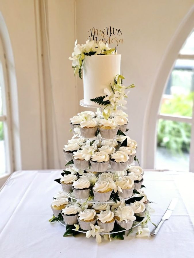 In questa foto che mostra Quale torta scegliere per il matrimonio" si vede una composizione di mini cupcake disposti a piramide, di colore bianco con crema a forma di rosa, accompagnati da decorazioni floreali. In cima alla struttura si trova una tortina cilindrica bianca decorata con fiori freschi e un cake topper dorato con la scritta 'Mr & Mrs Mendoza'