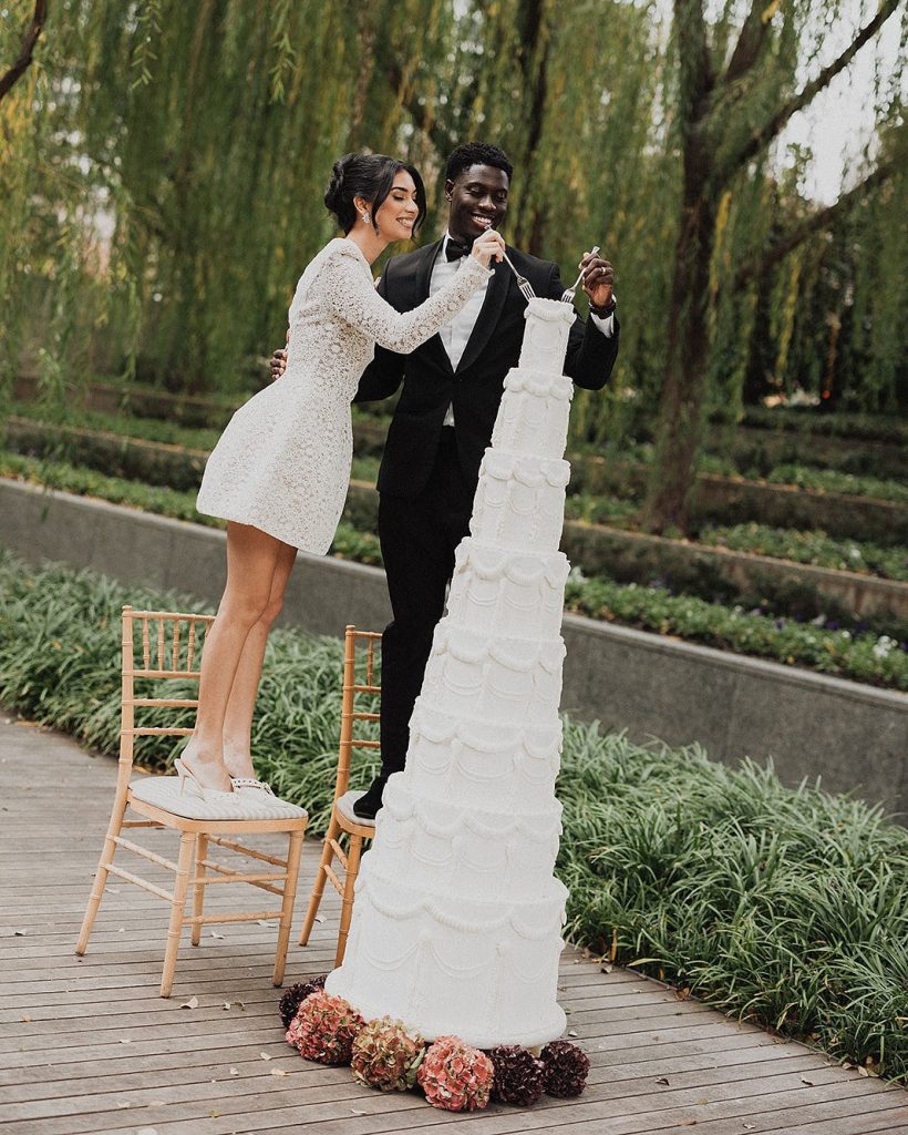 In questa foto che mostra Quale torta scegliere per il matrimonio" si vede una coppia di sposi mentre taglia una torta nuziale altissima di colore bianco. La torta è composta da numerosi piani impilati verticalmente e decorata con motivi eleganti a rilievo. Alla base, ci sono fiori secchi di colore rosa, rosso e bordeaux. La sposa, con un abito corto bianco e ricamato, è in piedi su una sedia per raggiungere la cima della torta, mentre lo sposo in smoking nero la sostiene e sorride. Lo sfondo mostra un giardino con alberi e aiuole verdi. L’immagine trasmette un momento giocoso e romantico.