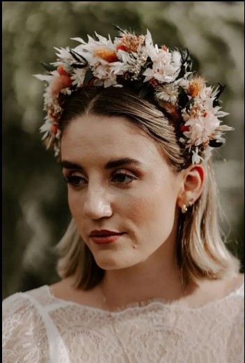In questa foto che mostra Acconciature sposa per capelli, si vede una sposa con capelli lisci di colore biondo chiaro, tagliati a caschetto. Indossa una vistosa corona floreale composta da fiori di colore bianco, rosa e arancio, con dettagli verdi e neri. Il look è completato da un abito da sposa in pizzo, visibile nella parte superiore. L’acconciatura è semplice ma impreziosita dalla corona, che dona un tocco boho-romantico. Lo sfondo naturale e sfocato crea un’atmosfera eterea e delicata