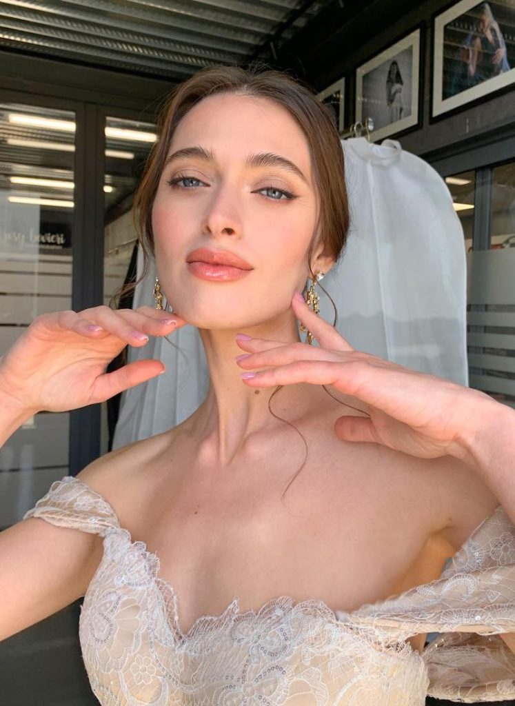 In questa foto, una modella posa con un abito da sposa in pizzo con scollo off-shoulder e un trucco sposa naturale nei toni del pesca