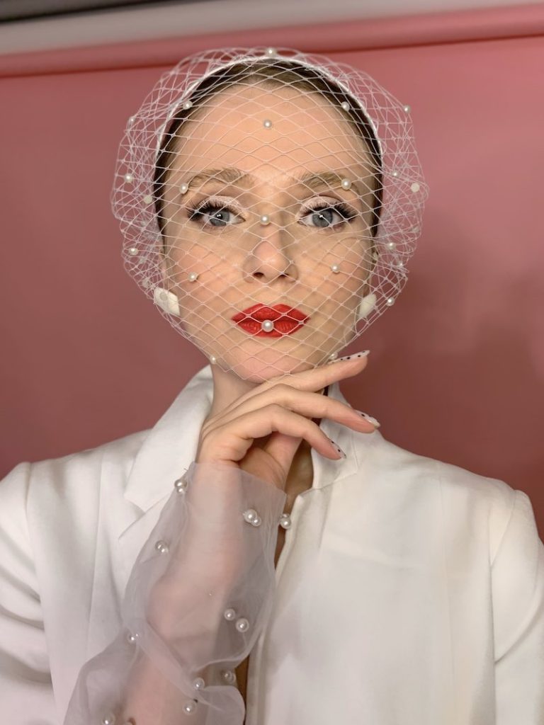 In questa foto, una modella posa con una veletta e un trucco sposa stile anni '50 con rossetto di colore rosso, base bianchissima e sguardo intenso firmato da Annartstyle. La modella indossa anche una veletta