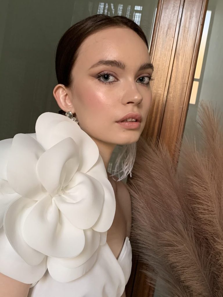 In questa foto, una modella posa con un abito da sposa con un grande fiore 3D e un trucco sposa naturale minimal