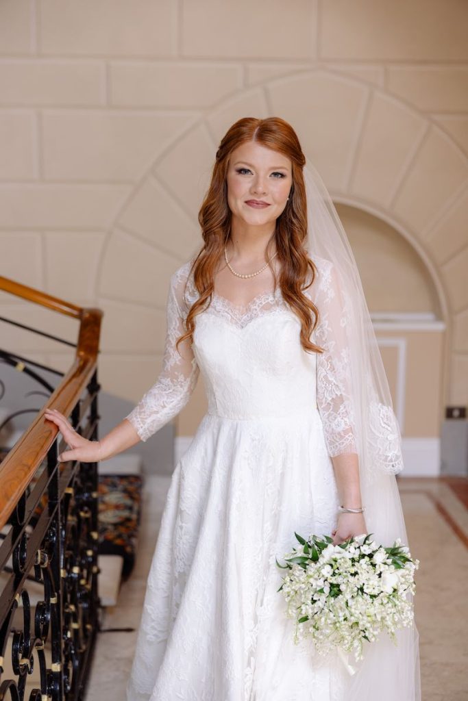 In questa foto, una sposa con i capelli di colore rosso e un trucco realizzato da Anna Ushakova e che punta l'attenzione sulle labbra, mentre scende le scale. La sposa indossa un abito con maniche di pizzo a tre quarti, dal taglio scivolato, un velo con bordo ricamato e porta tra le mani un bouquet di mughetto