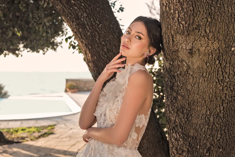 In questa foto, una modella posa appoggiata ad un albero e indossa un abito da sposa con un corpetto ricamato e un trucco naturale realizzato da Anna Ushakova