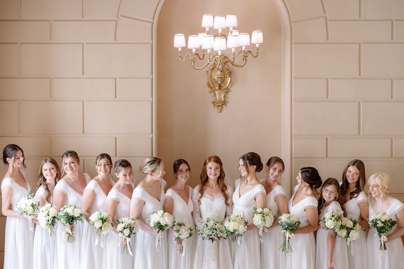 In questa foto, una sposa posa insieme alle sue 13 damigelle tutte vestite di bianco con bouquet di rose di colore bianco
