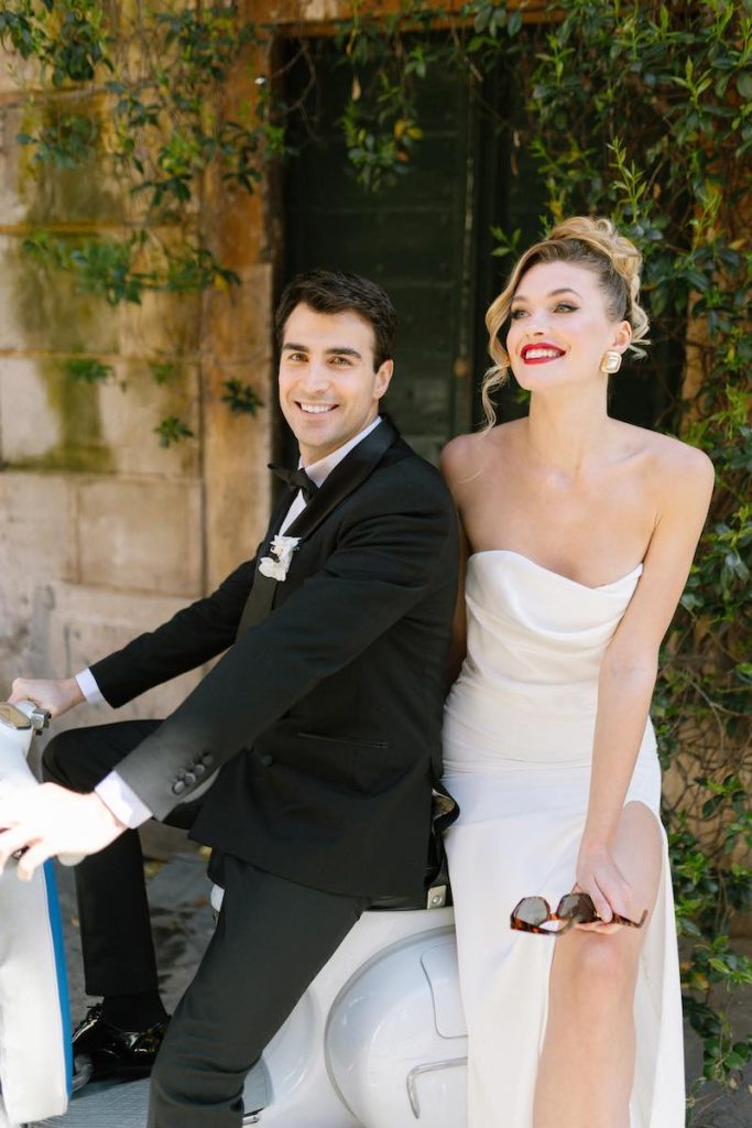 In questa foto, due sposi su una moto Vespa. Lo sposo indossa uno smoking mentre la sposa un abito a sirena senza spalline e un trucco deciso con rossetto di colore rosso 