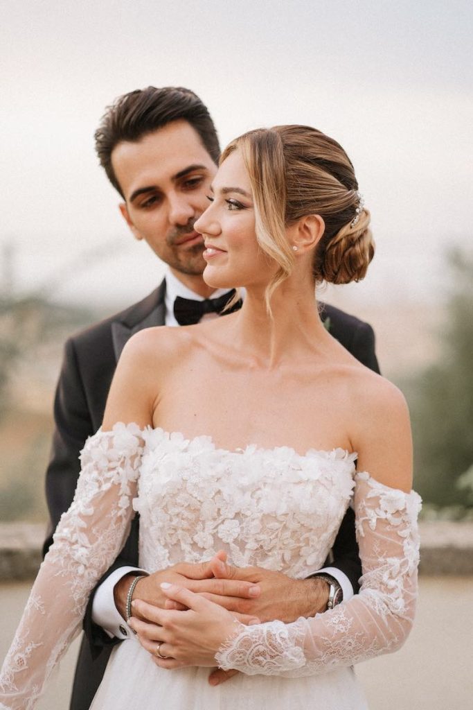 In questa foto, due sposi abbracciati l'uno davanti all'altro. La sposa  indossa un abito in pizzo floreale con manicotti e presenta un Beauty Look elegante e romantico realizzato da Annartstyle