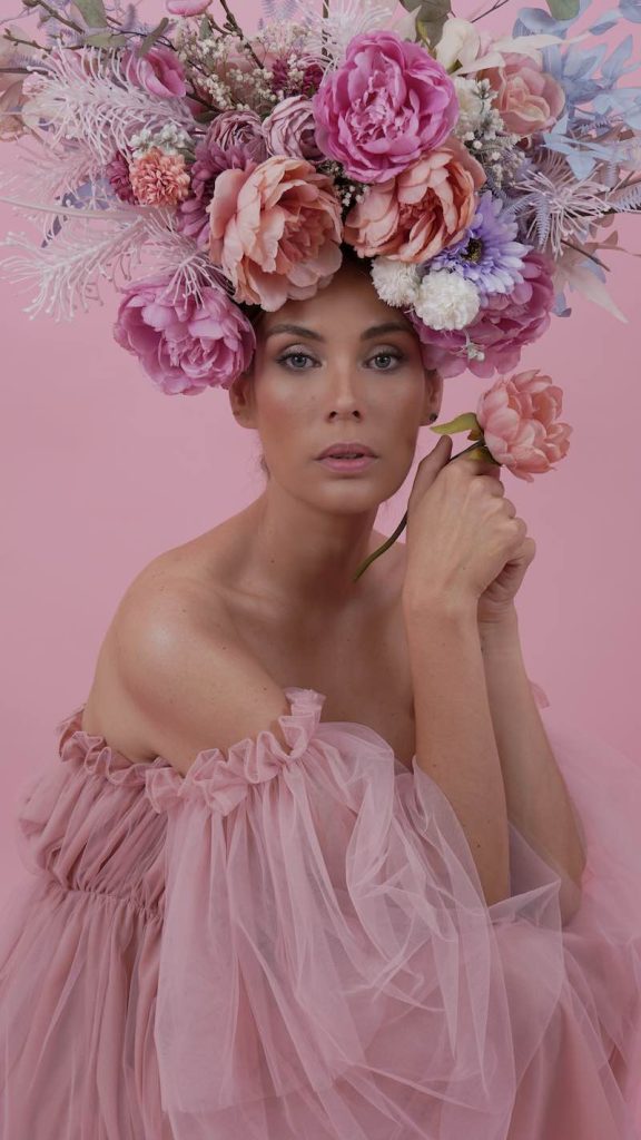 In questa foto, una modella posa su uno sfondo rosa, con un'acconciatura di fiori colorati in toni di rosa e lilla, indossa un abito di tulle di colore rosa. Anche il trucco realizzato da Anna Ushakova segue i toni del rosa
