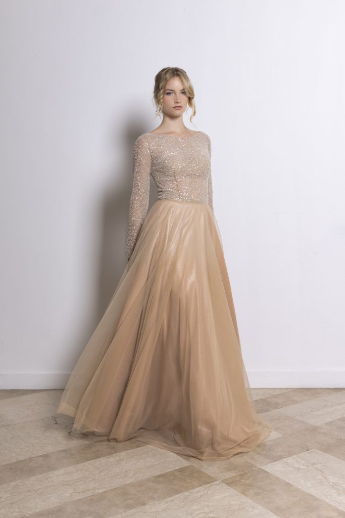 Una modella con capelli biondi raccolti in uno chignon morbido indossa un elegante abito da sera color champagne. Il corpetto è aderente, a maniche lunghe e completamente ricoperto di paillettes e microglitter, che creano un effetto luminoso e sofisticato. Lo scollo è leggermente a barca, mettendo in risalto il collo e le spalle. La gonna è ampia e vaporosa, realizzata in tulle leggero e semitrasparente con tonalità dorate calde e sfumature lucenti. Il tessuto fluttua verso terra in pieghe morbide, regalando movimento e leggerezza all’intero look. La modella è in piedi davanti a una parete bianca e un pavimento chiaro a piastrelle.