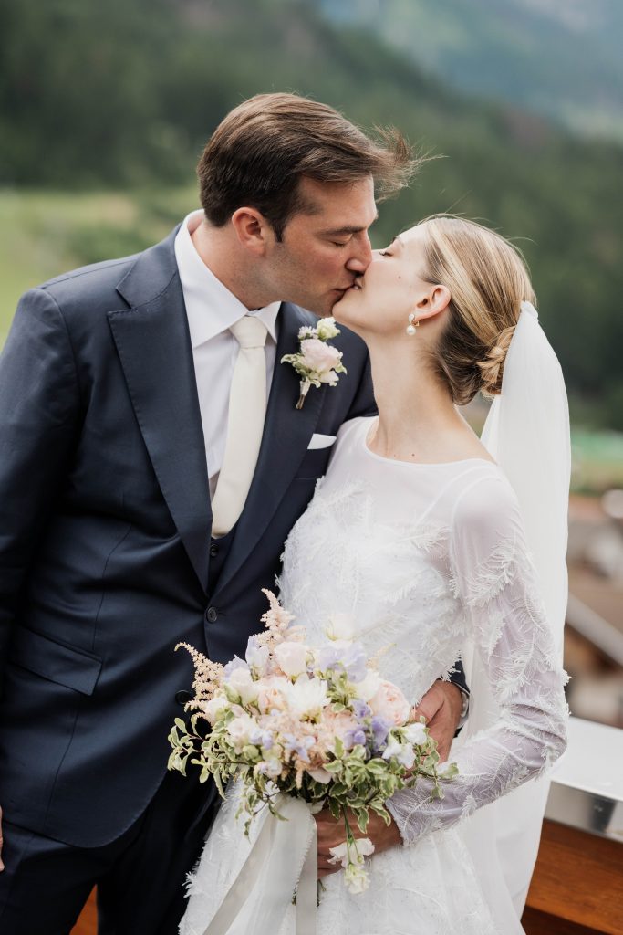 In questa fotografia romantica, una coppia di sposi si bacia teneramente all'aperto, con alle spalle un paesaggio montano sfocato e verdeggiante. Lo sposo indossa un elegante abito blu scuro con camicia bianca, cravatta color avorio e una boutonnière con rosa chiara appuntata sulla giacca. La sposa ha i capelli biondi raccolti in uno chignon basso e indossa un velo bianco fissato sul retro della testa. Il suo abito da sposa è bianco, a maniche lunghe, decorato con delicati motivi in rilievo tono su tono. Tiene in mano un bouquet romantico di fiori dai toni pastello – rosa cipria, lilla e bianco – con rami verdi e un nastro di seta bianco. La scena trasmette intimità, amore e serenità
