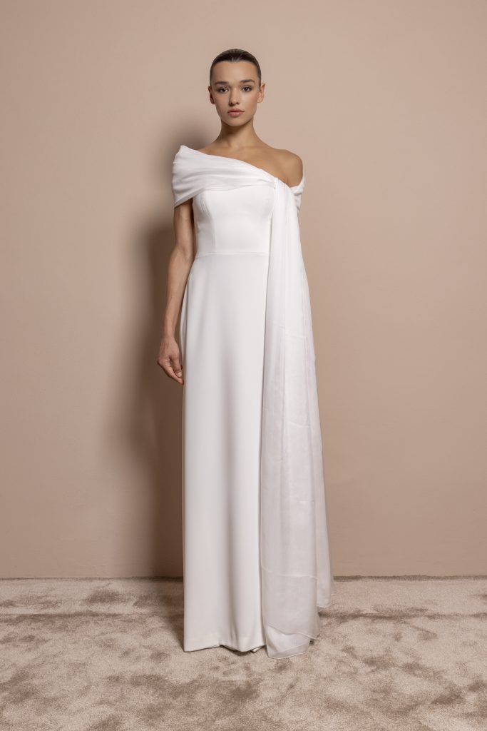 Una modella posa frontalmente, in piedi su un morbido tappeto beige chiaro, davanti a una parete rosa cipria. Indossa un abito da sposa lungo e liscio, in tessuto bianco. Il vestito ha una linea essenziale e scivolata, con taglio dritto e vita leggermente segnata. La particolarità è la spalla scoperta: un drappo di tessuto leggero è avvolto attorno al busto e ricade elegantemente su un lato, creando un effetto monospalla raffinato. La modella ha i capelli raccolti e un’espressione serena e composta.