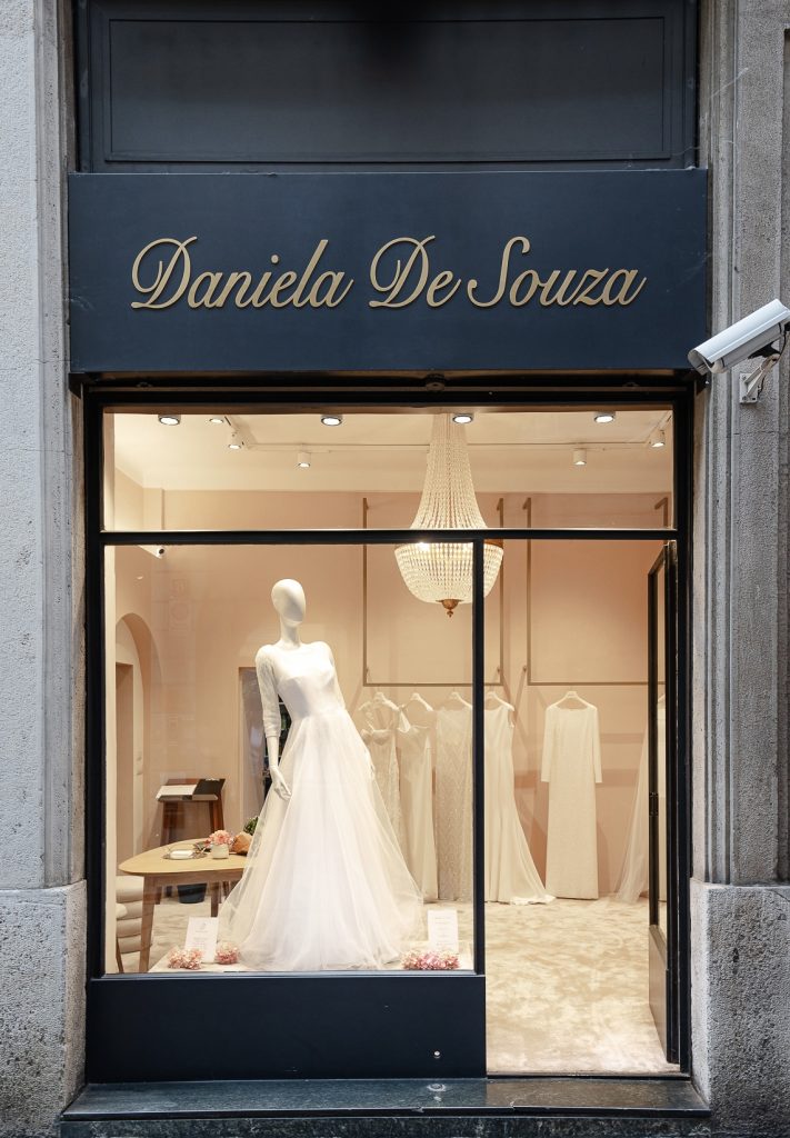 Vista frontale della vetrina dell'atelier Daniela De Souza. In primo piano di vede un abito da sposa con un'ampia gonna in tulle color bianco ottico. 
Un lampadario antico con cristalli e dietro una rella con alcuni abiti da cerimonia appesi