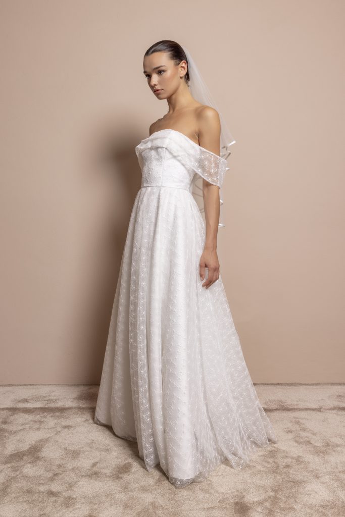 Una modella è in posa di tre quarti, rivolta verso sinistra, su un tappeto beige chiaro davanti a una parete rosa cipria. Indossa un abito da sposa lungo, bianco, con un corpino rigido e spalle scoperte. Il vestito ha maniche calate che avvolgono le braccia delicatamente e una gonna ampia che scende fluida fino al pavimento. Il tessuto è decorato con un delicato motivo floreale in pizzo. La modella porta un velo bianco fissato ai capelli raccolti in uno chignon basso. L’espressione è elegante e composta.