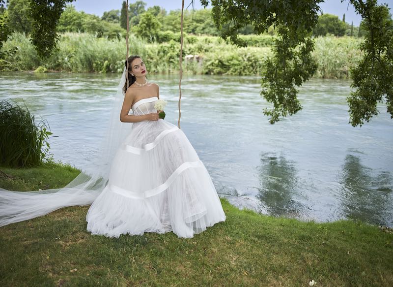 In questa foto, una modella posa su un'altalena a bordo lago con un abito da sposa ampio in tulle, con balze e corpetto steccato della collezione sposa Elisabetta Polignano 2026