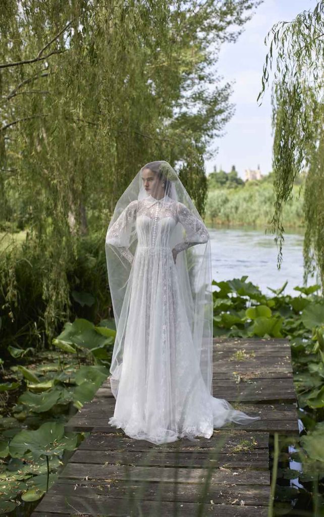 In questa foto, una modella posa su un piccolo ponte sospeso su un lago con un abito da sposa scivolato in pizzo con maniche balloon e colletto della collezione sposa Elisabetta Polignano 2026