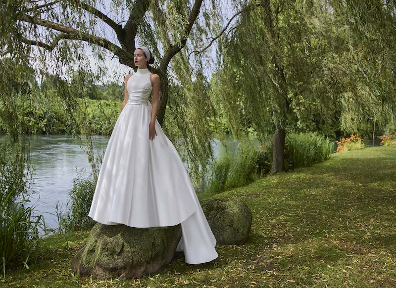 In questa foto, una modella posa su un albero con un abito da sposa ad A con scollo halter e lungo strascico collezione sposa Elisabetta Polignano 2026